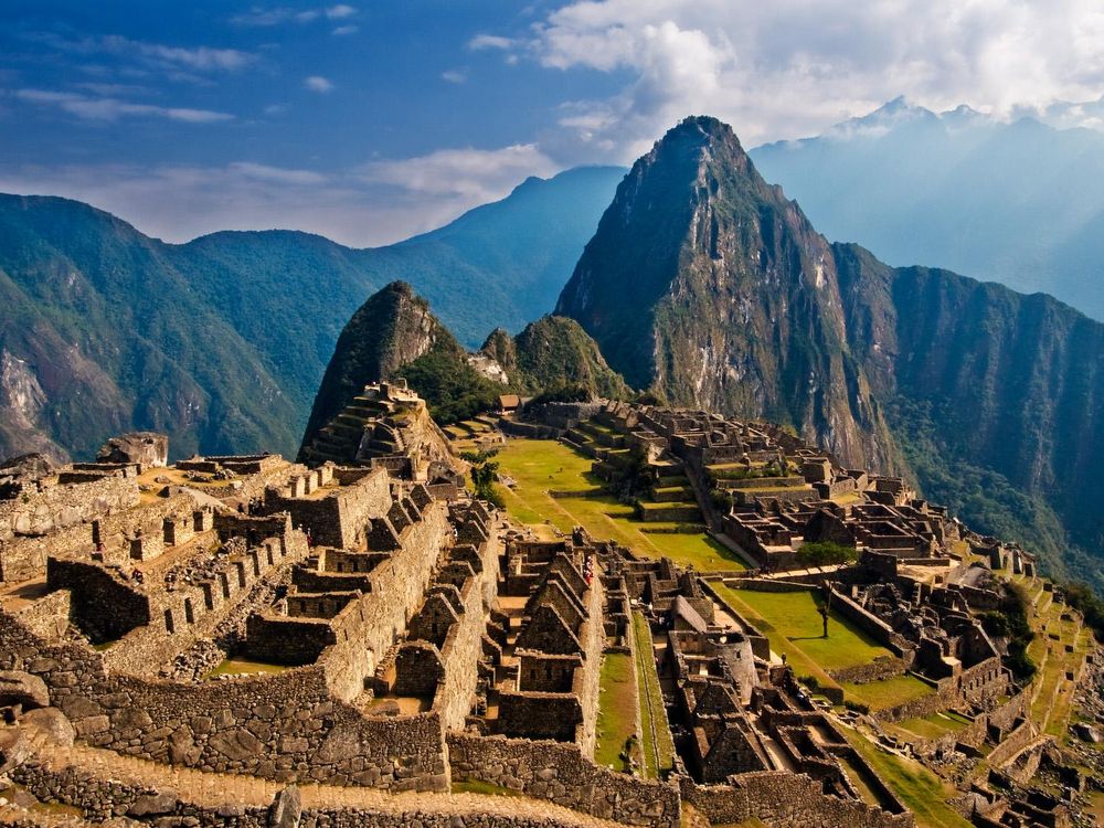 An aerial view of Machu Picchu at sunrise, with an orange light coloring the 15th century stone structures and a far off peak covered in green foliage