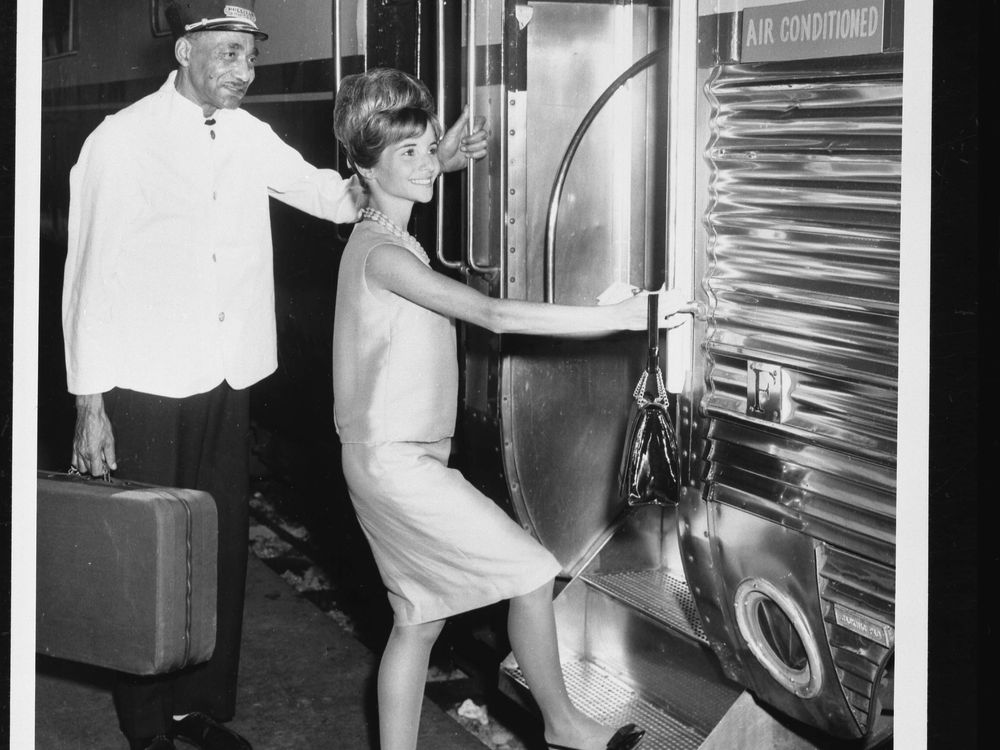 A black-and-white photograph of an African American male porter holding a suitcase while a white woman steps up into a rail car.