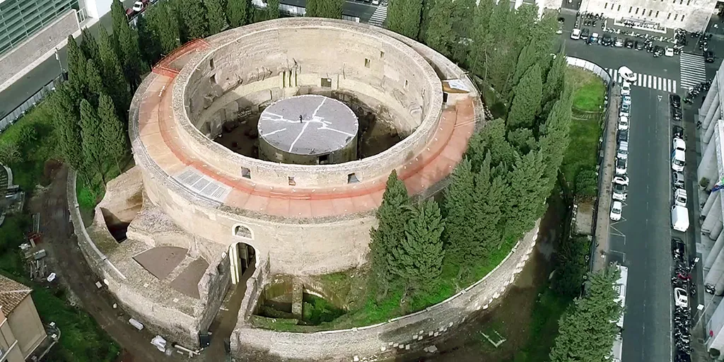 An aerial shot of a large circular building, with reddish and yellow stones, surrounded by trees on all sides