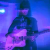A woman with long dark hair and dark outfit plays a white electric guitar on a dimly blue lit stage. She and her guitar are slightly blurry.