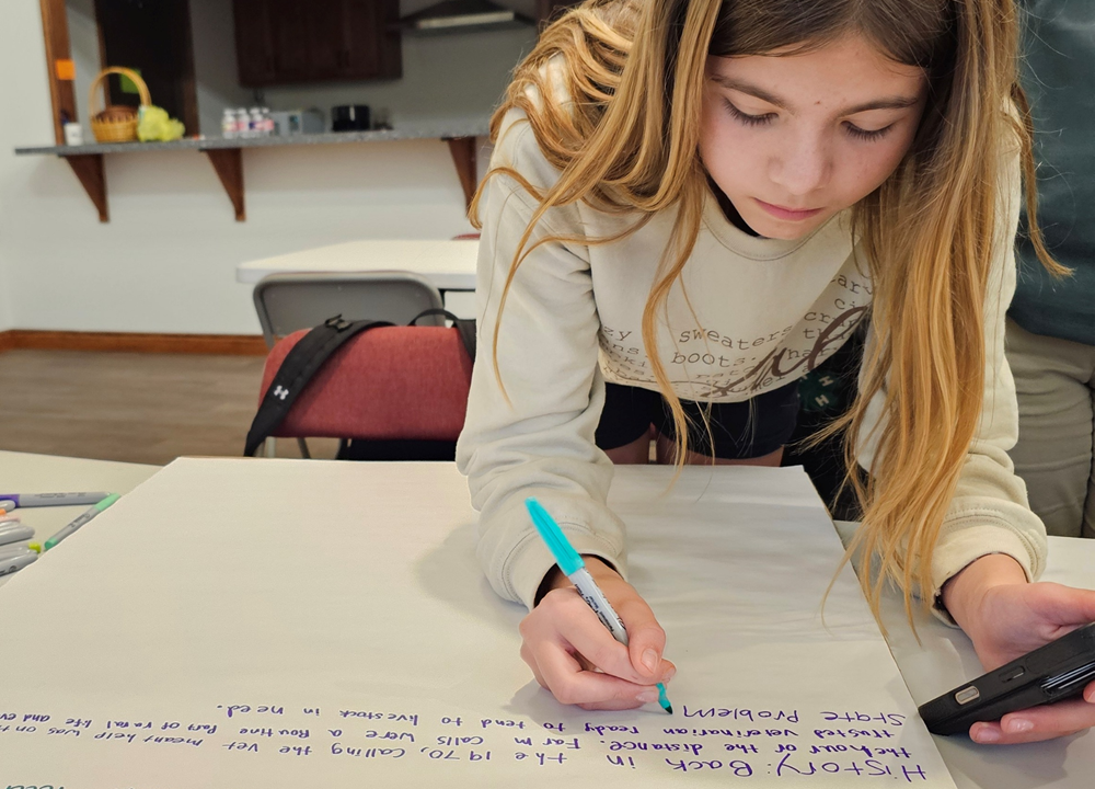 A student leans over a table, writing on a large poster board with a marker during a classroom activity. Surrounding the workspace are colorful pens, papers, and notes, while additional tables, chairs, and a kitchen area with cabinets and a counter appear