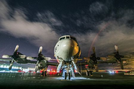 A massive aircraft with four propellers sits on a wet tarmac reflecting the bright red and white lights of the surrounding air base. The night sky above is cloudy, though some bright stars are visible.