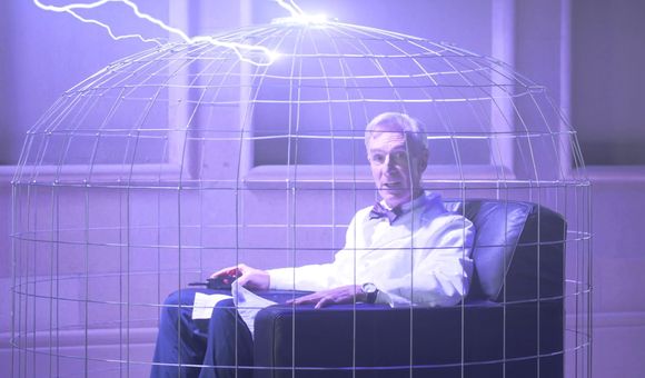 A man wearing a lab coat sits in a chair inside a wire cage with electric bolts coming down