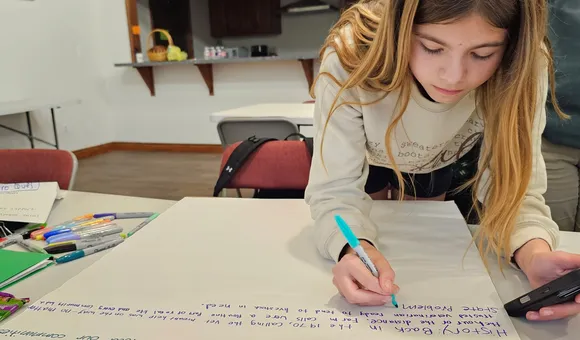 A student leans over a table, writing on a large poster board with a marker during a classroom activity. Surrounding the workspace are colorful pens, papers, and notes, while additional tables, chairs, and a kitchen area with cabinets and a counter appear