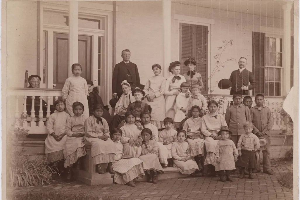A sepia toned image of a group of young women, with a few young boys and girls, sitting in front of a white house on a porch. Pratt stands in dark garb behind the group
