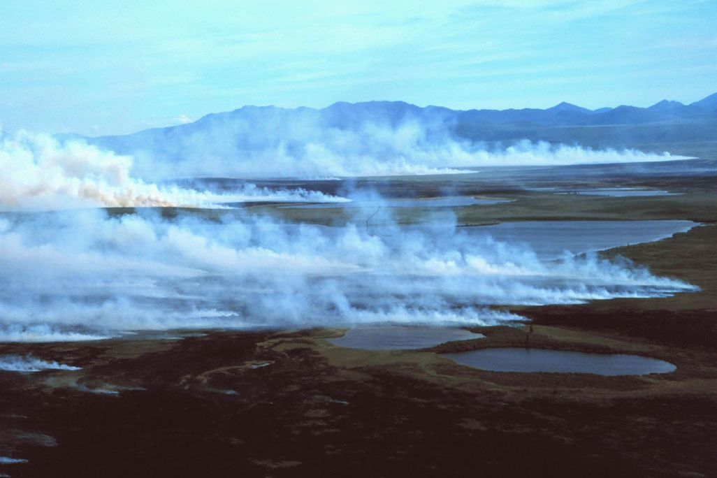A photograph of a tundra landscape with mountains on the horizon and smoke rising from the ground