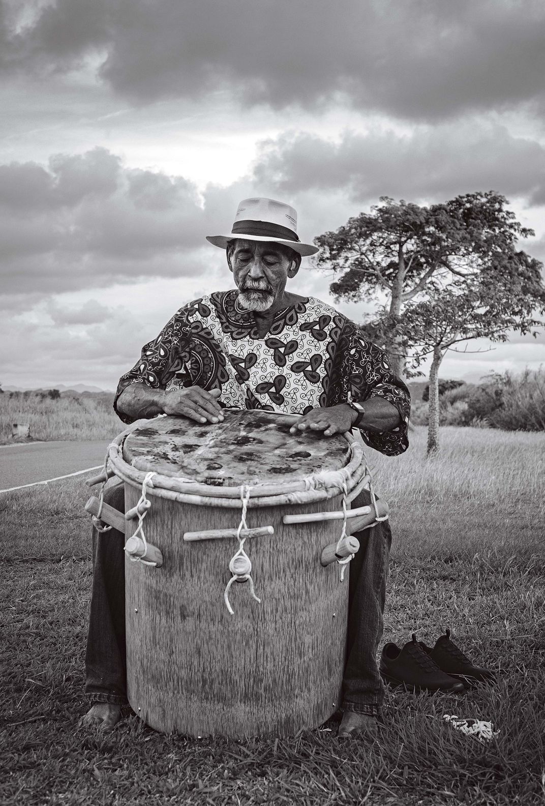 a black and white photograph of a man playing a  drum made out of goatskin and a palm tree trunk