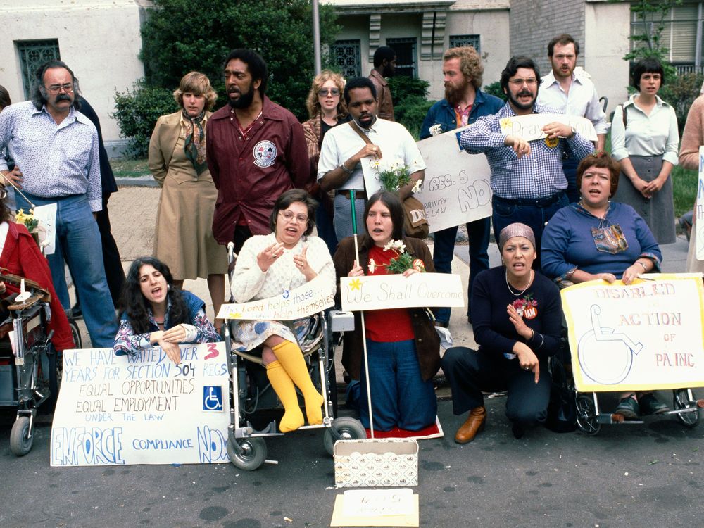 A color photograph of a group of protesters, including Judy Heumann, who is wearing bright yellow stockings. One sign reads: "We have waited over 3 years for Section 504 Regs...ENFORCE compliance NOW"