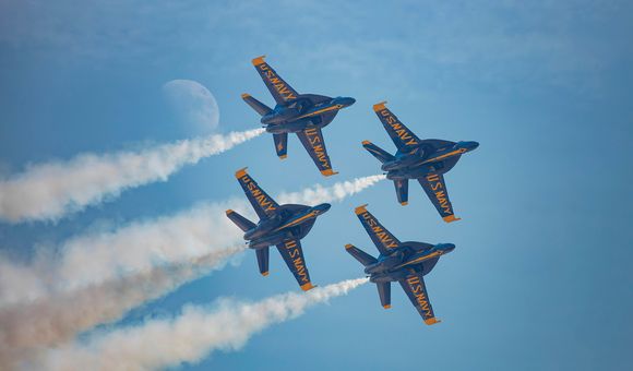 Four military jets fly in a diamond formation across a blue sky. On the underside of each jet's wings, "US Navy" is painted in bold yellow letters. White airshow smoke streams from each jet. The moon is visible in the sky.