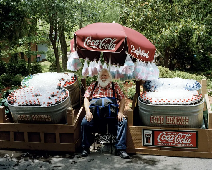 Santa dressed in regular clothes sitting next to a Coca Cola stand