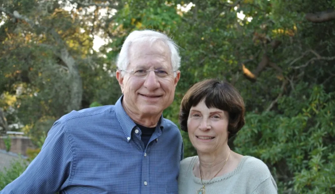 Portrait of a man and woman standing outside in front of green trees.