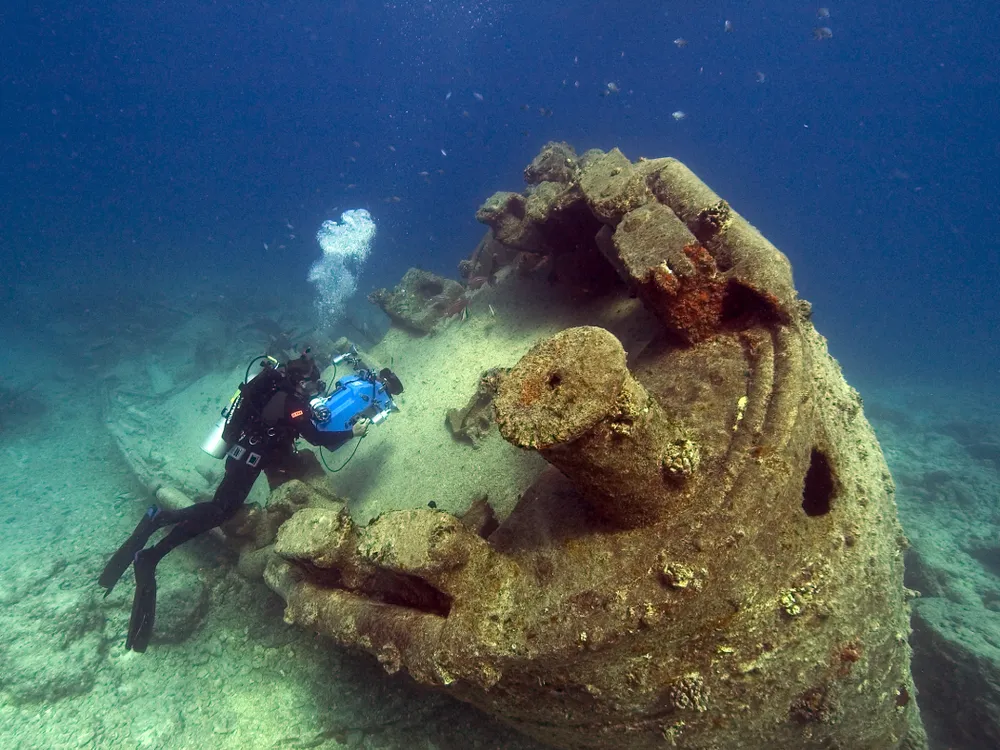 A diver uses a camera to study a barnacle-covered submarine resting on the seafloor
