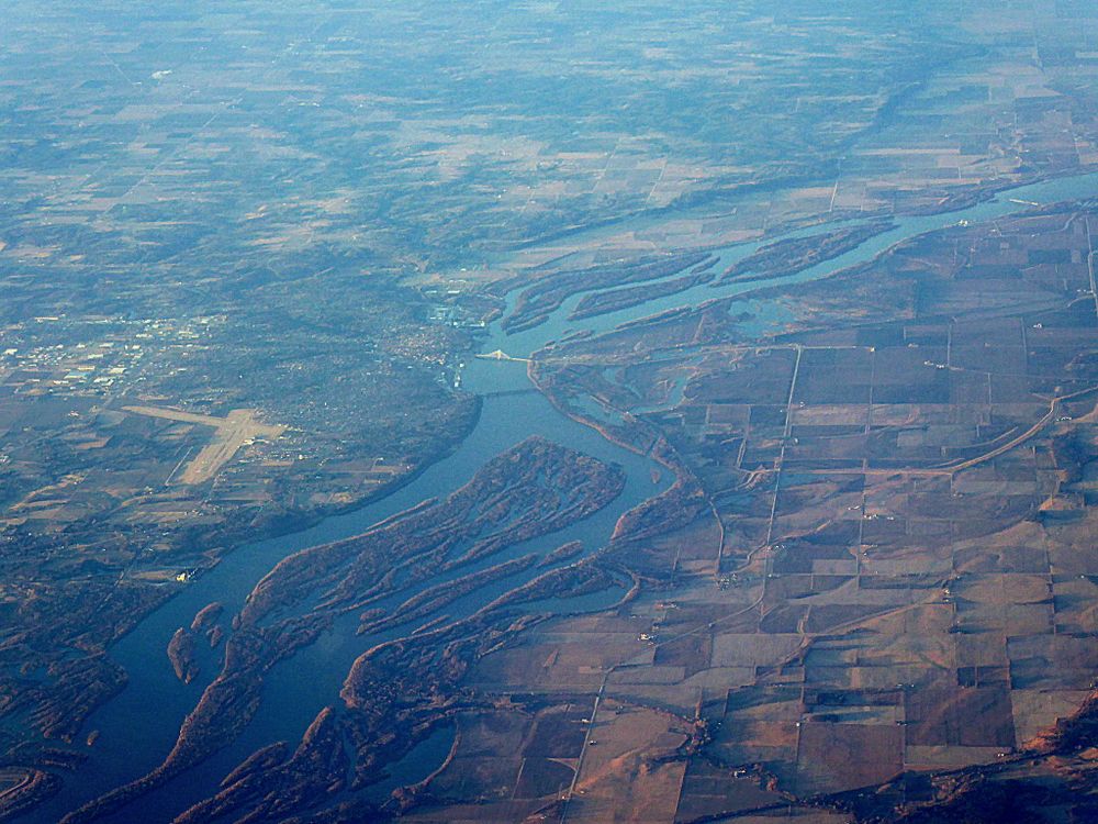 An aerial photo of the Mississippi River flowing through Iowa. The river runs through the middle of the photo, separating patches of fields on the right and more urban and suburban areas on the left. 