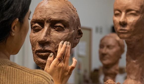 A person sculpts a clay bust. Two additional sculptures appear in the background.