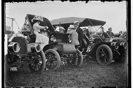 A black and white photograph featuring cars with people conversing.