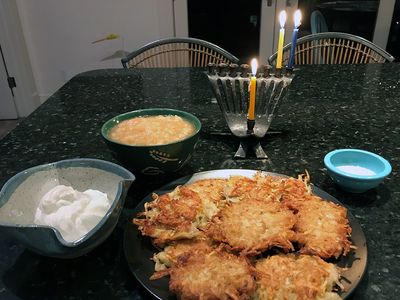 A plate full of golden brown potato pancakes are placed on a dining table. Behind them are bowls of sour cream, apple sauce, salt, and a menorah with three candles lit.