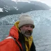 A man wearing an orange rain jacket sits on a boat, framed by the ocean and a glacier behind him.