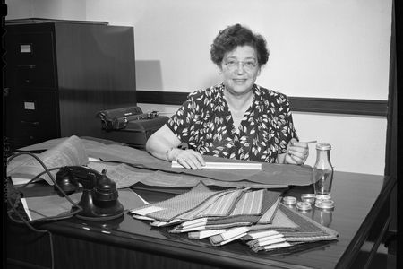 Black and white photograph of a woman sitting behind a desk. She has short, wavy hair and wears glasses. The desk is covered in rectangular carpet samples and blueprints are spread across the desk. A black telephone rests on the desk amid the supplies.