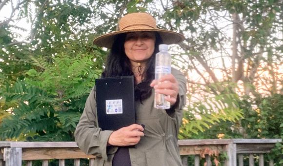 A woman wearing a straw sun hat stands on a dock with a forest behind her, holding out a plastic water bottle and smiling.