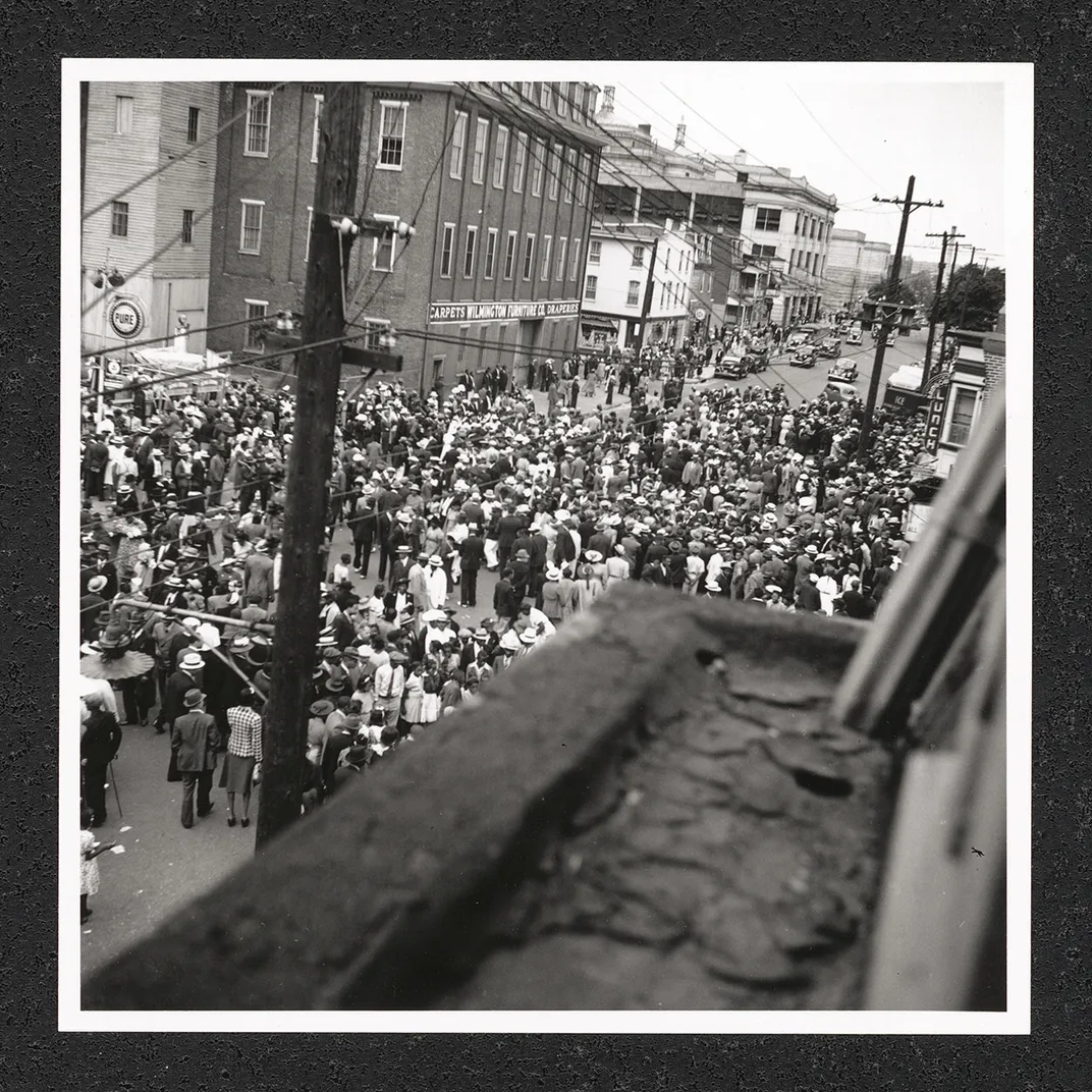 From above, a dense crowd of people in the streets. The corner of a rooftop is out of focus in the foreground. Black-and-white photo.