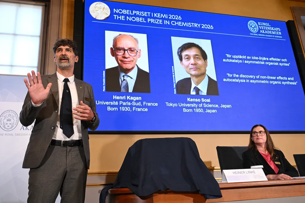 A man in a suit and tie standing in front of a large screen with pictures of two other men and words, with a woman sitting at a table nearby