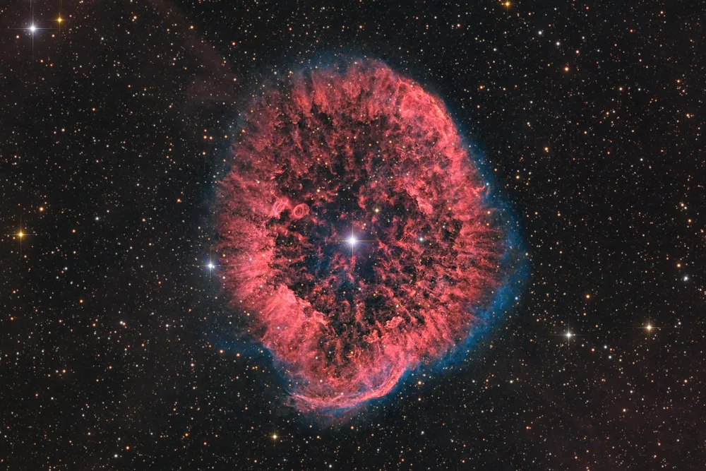 a red billowing ring of cosmic gas and dust surrounds a bright star with other stars appearing as several pinpricks of light in the background