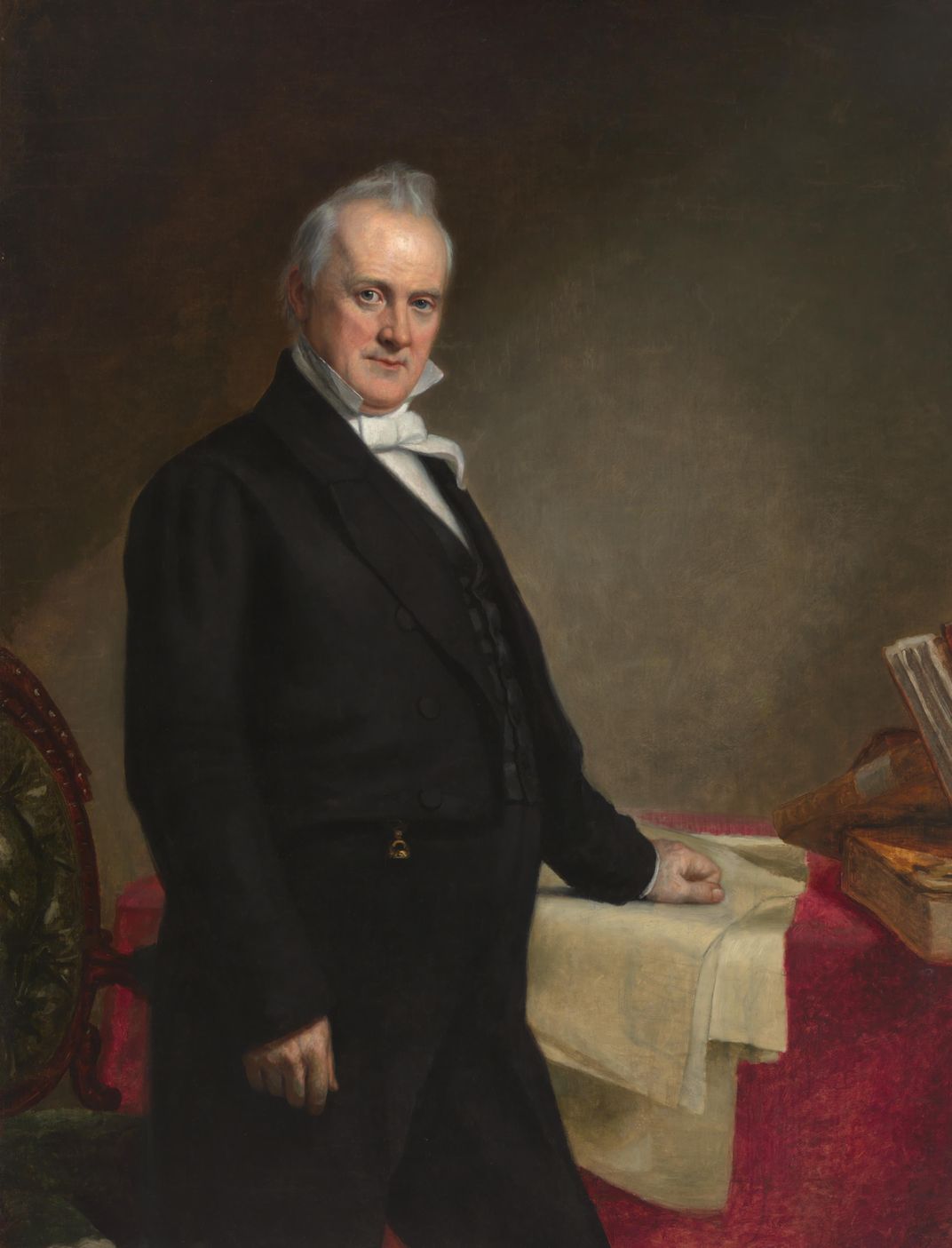 A white man in a suit, with gray hair, stands in front of a red table and chair and faces the viewer