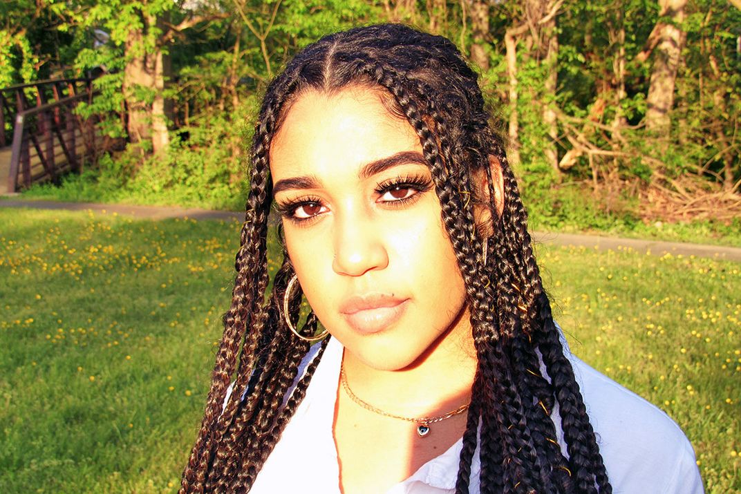 Portrait of a woman standing in a park. She has long dark braids and gold hoops.