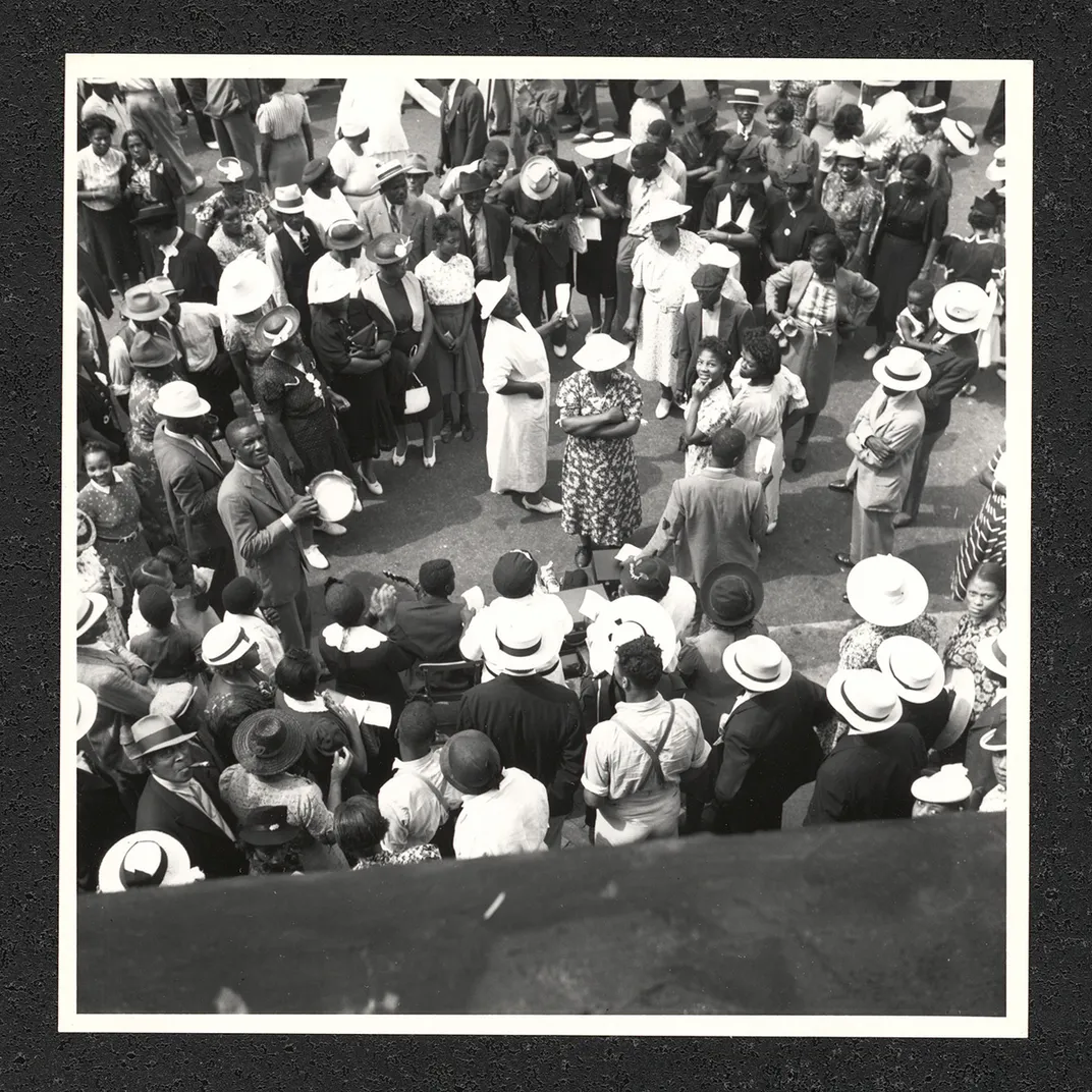 From above, a crowd of people form a circle outdoors. Some holds musical instruments, like a tambourine. Black-and-white photo.