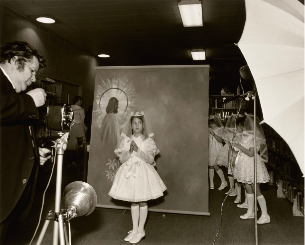 Black and white photograph of a girl posing for a first communion picture. A dropdown background image of a man with a halo around his head. A photographer stands with a camera in front of the girl.