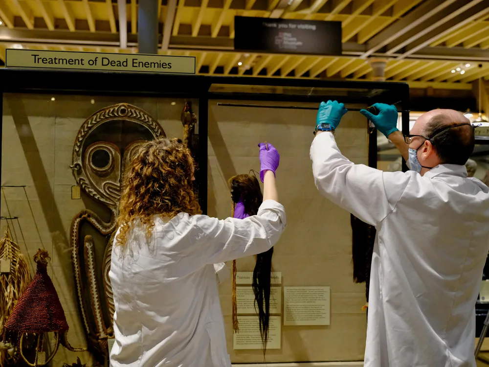 Two people in white lab coats and gloves; left, person with curly hair, and right, person with short balding hair, are in the process of removing tsantsa, small fist-sized skulls with long hair, from the display case