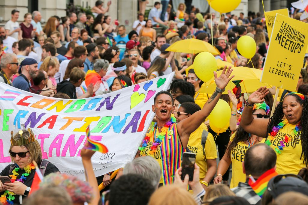 People in bright yellow shirts, including one person smiling with their arm raised, boast rainbow flags and march in support of LGBTQ people