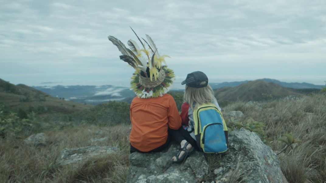 Film still of two kids sitting on a rock, facing away toward a mountainous landscape. One wears a feathered headdress and the other a backpack.