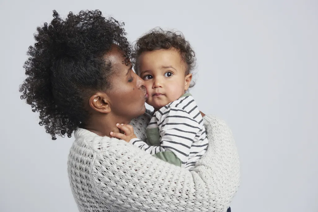 A mom holds her toddler and gives him a kiss on the cheek against a grey background