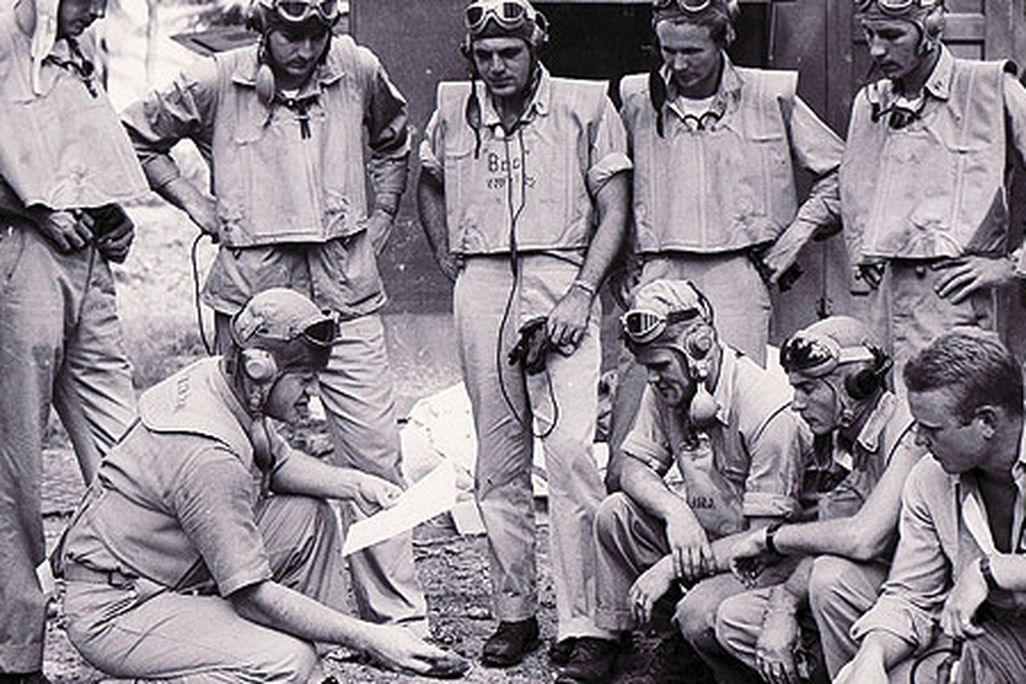 At the Black Sheep Squadron's base on the South Pacific island of Espiritu Santo, Boyington (holding paper) briefs his pilots on an upcoming mission.
