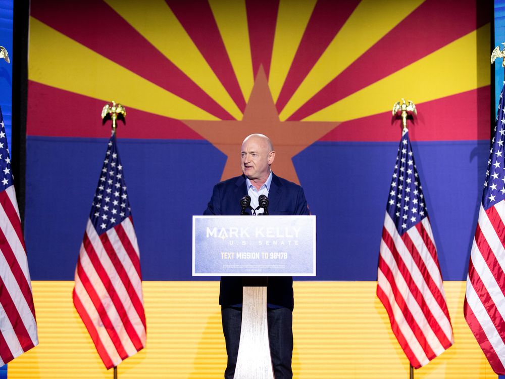 Mark Kelly stands on stage at a podium in front of the Arizona flag 