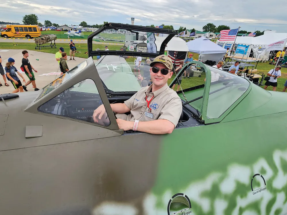 A white man in his 30s sits in the cockpit of a World War II-era military airplane. He is smiling and wearing sunglasses and a ballcap.