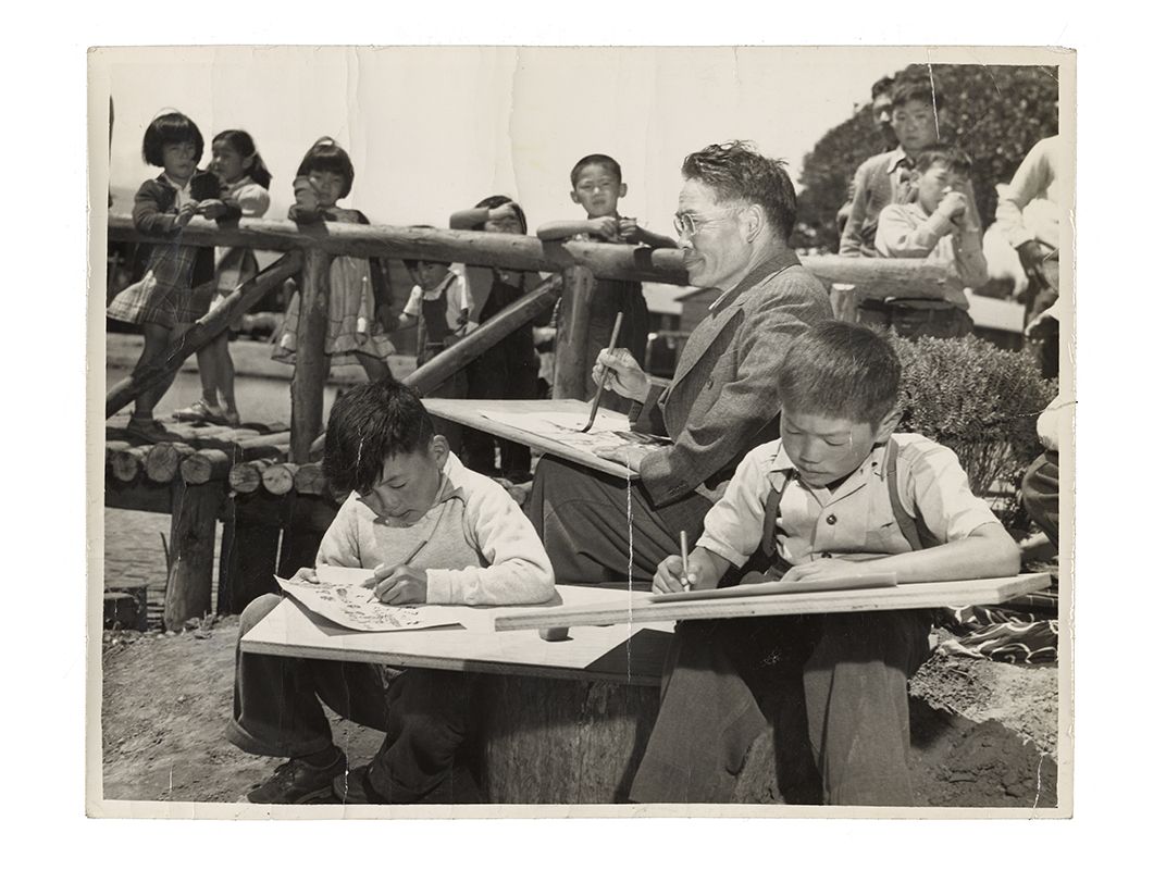 Grayscale image of an adult man teaching a children's art class while other children on a bridge in the background watch.