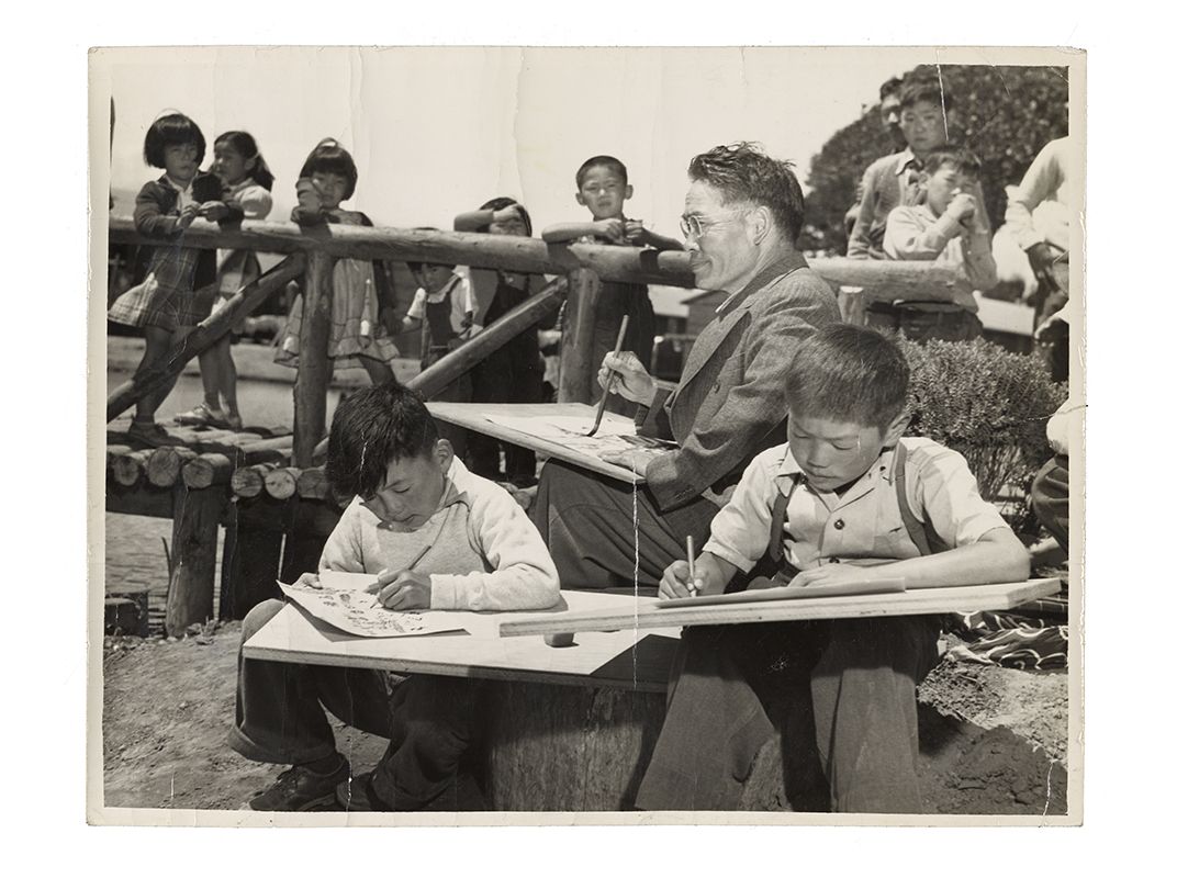 Gray scale image of adult man teaching a children’s art class while other children on a bridge in the background look on.