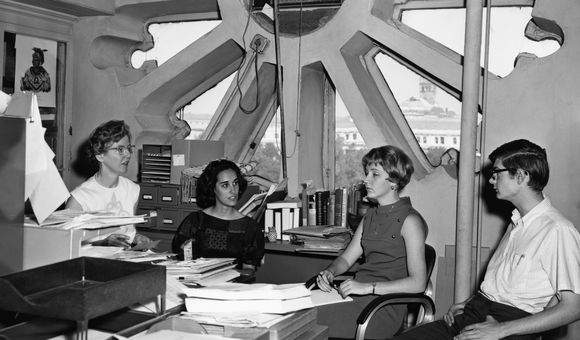 In a black and white photo, three women and one man sit together in a room with a large circular window behind them, surrounded by papers.