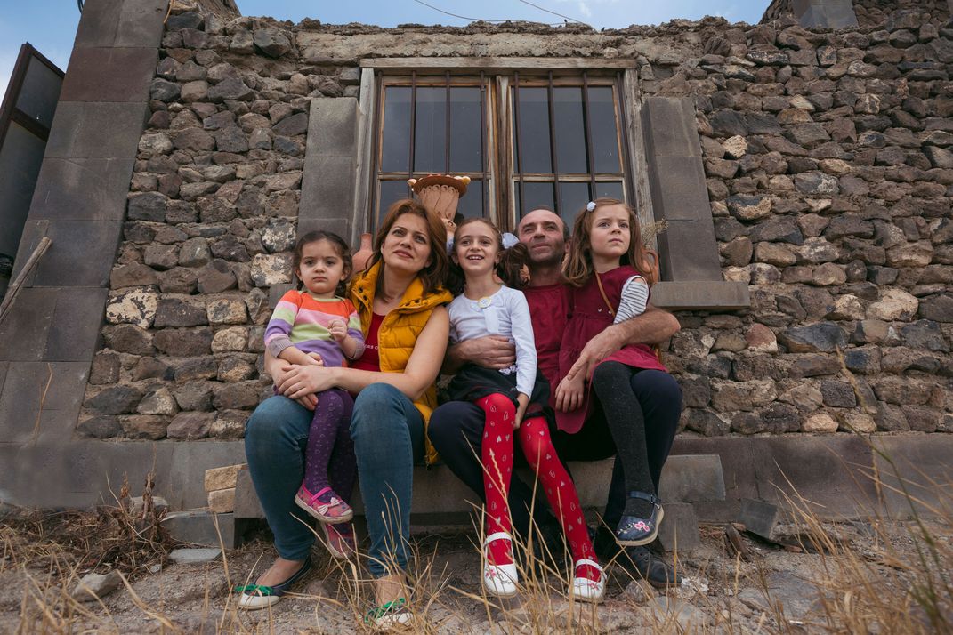 A man and his wife sit in front of a stone wall, their three young daughters sitting in their laps.