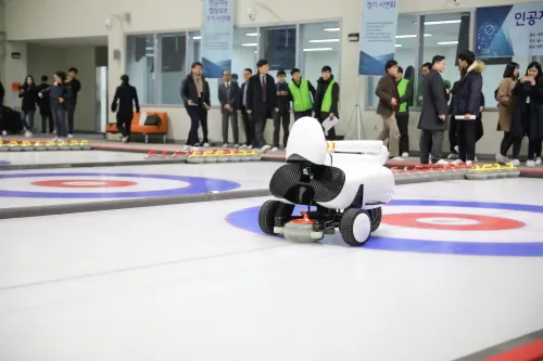 A white robot with a teardrop-shaped "head" appears to crouch down, rolling on wheels, as it pushes a round curling stone forward over ice; many onlookers watching