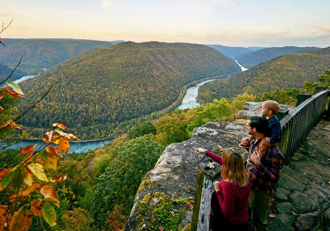 Visitors stand along the stone overlook at Grandview, looking out across the New River Gorge. The river winds through forested mountains below, with early autumn color visible across the landscape.