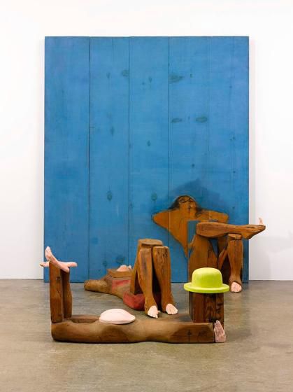 Wooden sculpture of three figures at the beach in various reclining positions. One figure wears a yellow hat and nothing else, one a red bikni, the other reads a book. Two  rest against a wooden panel painted blue. 