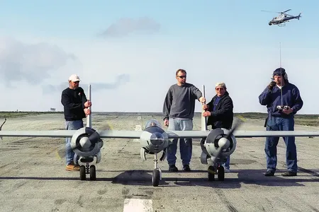 Four men stand on a paved runway behind a large, grey twin-engine remote-controlled model airplane. The man on the far right holds a radio transmitter with a long antenna, while a small helicopter is visible in the sky behind him. The scene is set under
