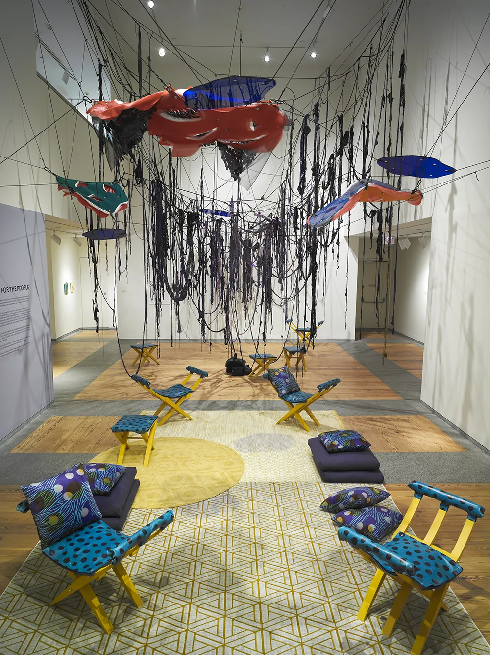 Gallery installation of blue and yellow patterned chairs, yellow patterned carpets, blue patterned pillows, and red, green, and blue hanging from the ceiling.