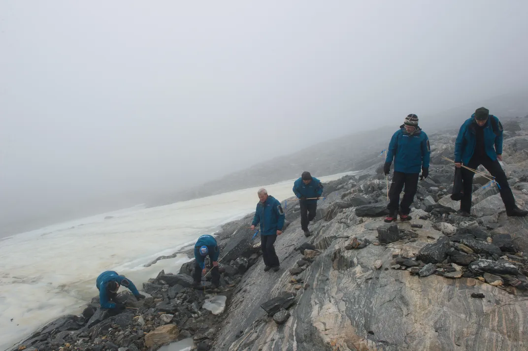 six people in blue jackets walk along rocks and examine the ground with ice in the background