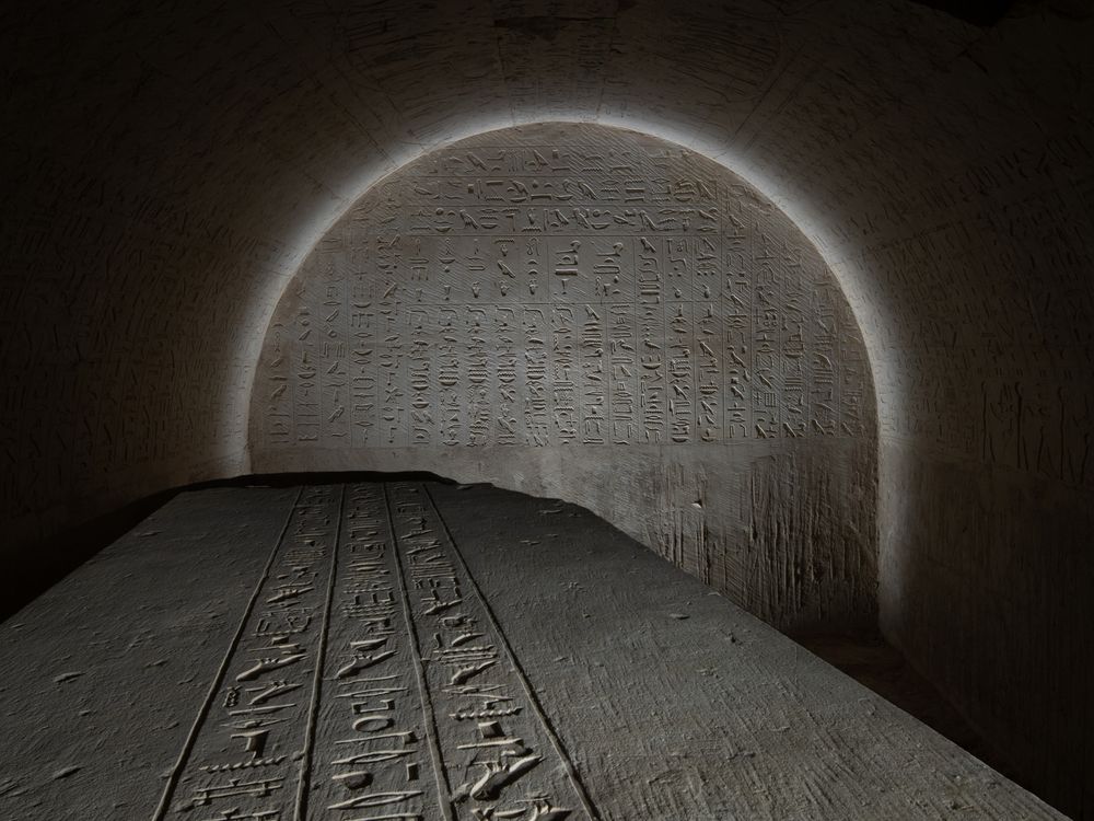 the top of a sarcophagus covered in writing with a glyph-covered wall behind it lit in an arch