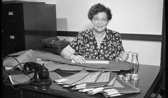 Black and white photograph of a woman sitting behind a desk. She has short, wavy hair and wears glasses. The desk is covered in rectangular carpet samples and blueprints are spread across the desk. A black telephone rests on the desk amid the supplies.