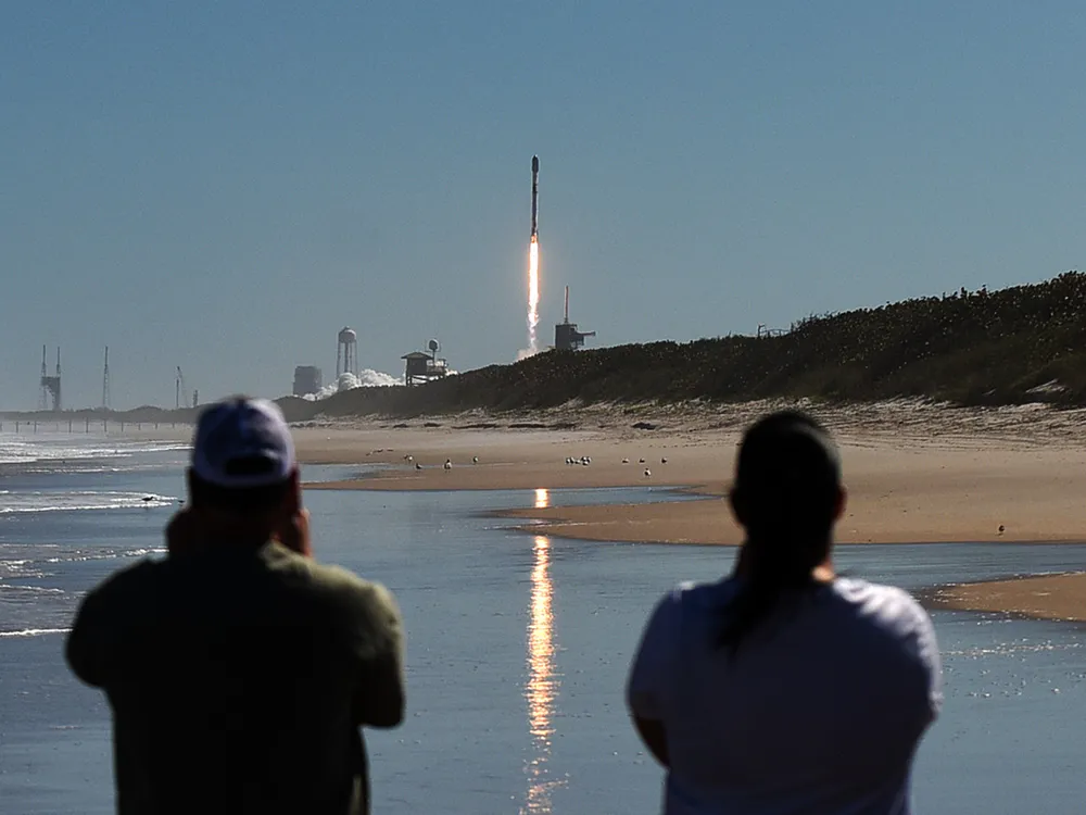 Image of SpaceX’s Falcon 9 rocket lifting off in Florida with blue sky and trail of fire, onlookers in foreground